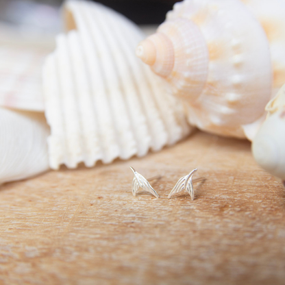 Silver mermaid tail-shaped earrings on a wooden surface with seashells in the background