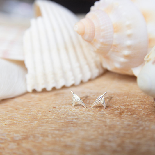 Silver mermaid tail-shaped earrings on a wooden surface with seashells in the background