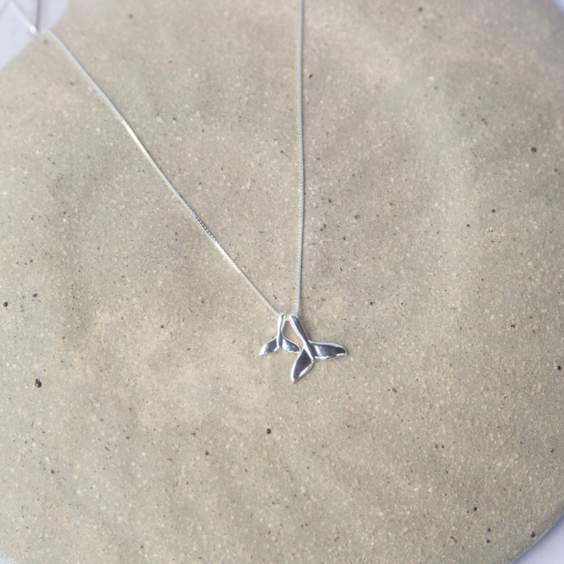 Silver whale tail necklace on a beige stone surface
