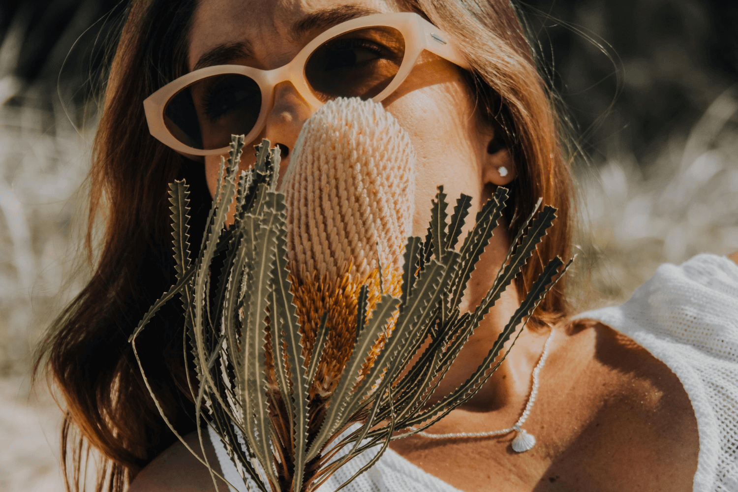 Woman wearing silver necklace holding a dried plant in front of her face with a blurred background